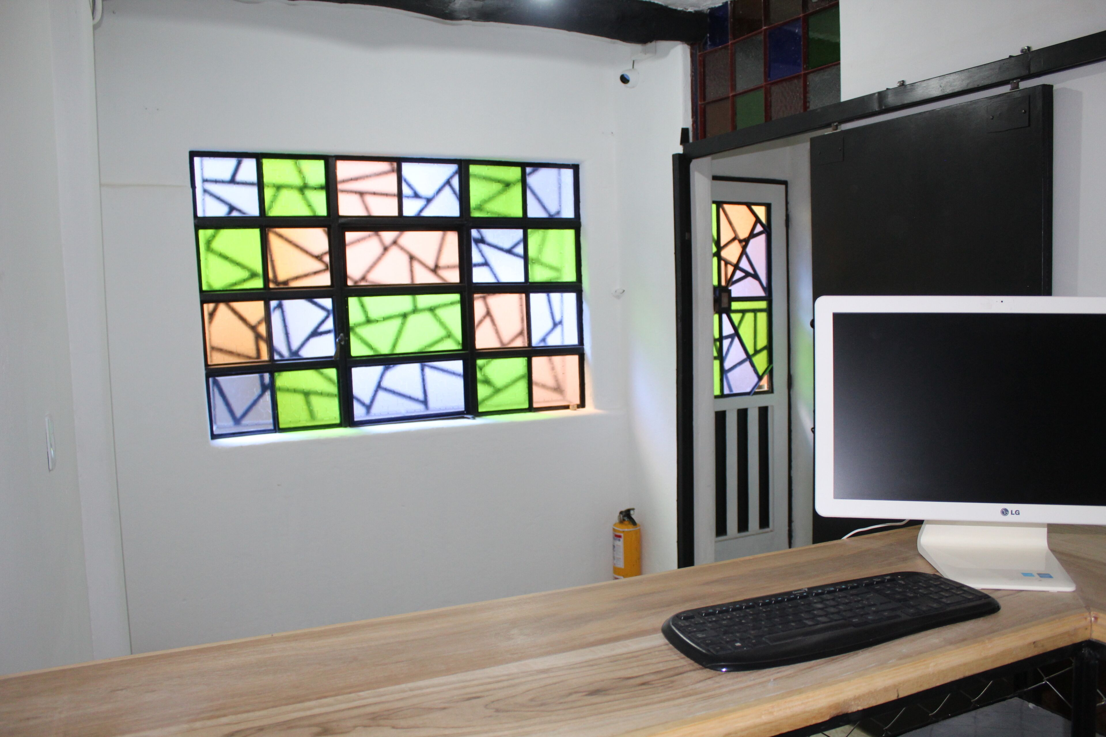 Una habitación con un escritorio de madera, un monitor de computadora y un teclado. En la pared hay una ventana de vidrio emplomado con colores.