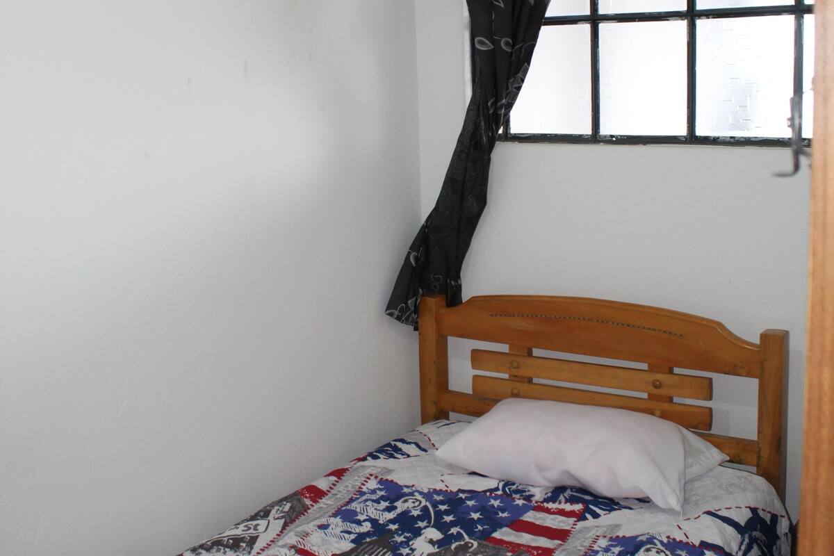 Ein kleines Zimmer mit einem Einzelbett, einem hölzernen Kopfteil, einer gemusterten Bettdecke und einem Fenster mit schwarzem Vorhang.