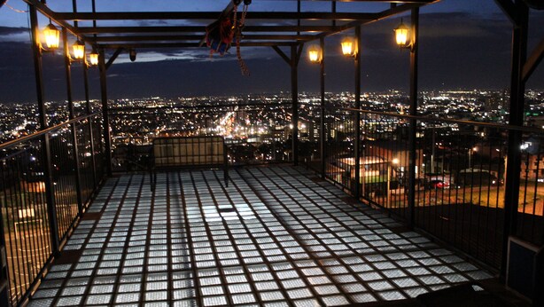 Eine Dachterrasse mit einem gitterförmigen Boden, Metallgeländern und Blick auf eine nächtliche Skyline.