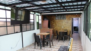 Ein Restaurant mit Fliesenboden, Holztischen und -stühlen, einem an der Wand befestigten Fernseher und einer Backsteinwand.