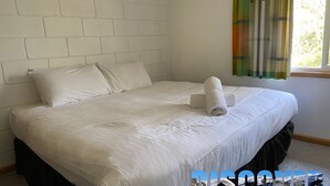 Ein ordentlich bezogenes Bett mit weißen Leinen und einem zusammengerollten Handtuch, eine schlichte weiße Ziegelwand und ein Fenster mit Vorhang.