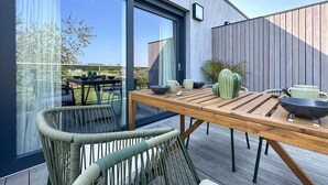 A modern outdoor dining area with a wooden table and chairs, a cactus plant, and a view of the surrounding landscape.