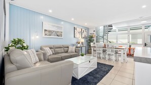 A modern living room with a beige sofa, a glass coffee table, and a dining area with white chairs and a wooden table.