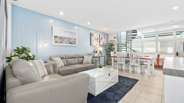 A modern living room with a beige sofa, a glass coffee table, and a dining area with white chairs and a wooden table.