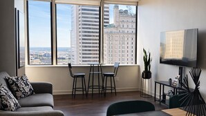 A modern living room with a sofa, dining table, chairs, a ceiling fan, a TV, and a large window with a city view. - Downtown Dallas - King Bed + Free Parking - Pool (Dallas)