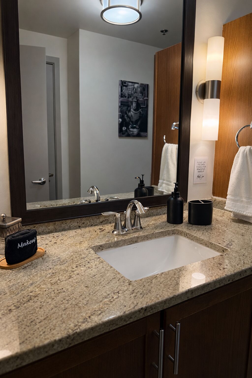 A bathroom with a granite countertop, a white sink, a large mirror, a towel rack, and a makeup bag.