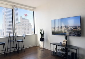 A modern living room with a large TV, a dining area with stools, a potted plant, and a view of a cityscape through a large window.