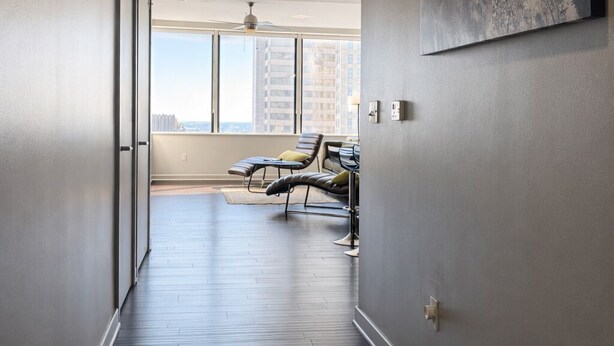 A hallway with dark wooden flooring, a ceiling fan, and a large window at the end leading to a seating area with chairs and a table. - Cozy Retreat with Rooftop Pool, Parking, and Spacious Comfort (Dallas)
