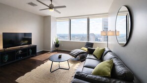 A modern living room with a flat-screen TV, a ceiling fan, a large window with a city view, a round mirror, and a leather sofa with cushions. - Cozy Retreat with Rooftop Pool, Parking, and Spacious Comfort (Dallas)