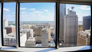Ein weiter Panoramablick vom Fenster eines Hochhauses auf eine Stadtlandschaft mit zahlreichen Gebäuden, darunter ein markanter Wolkenkratzer mit einer roten Skulptur auf dem Dach.
