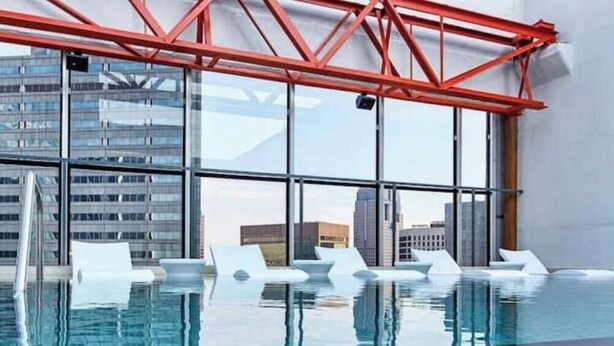 Ein moderner, überdachter Pool mit großen Glaswänden, weißen Liegestühlen und einer roten Metallkonstruktion darüber.