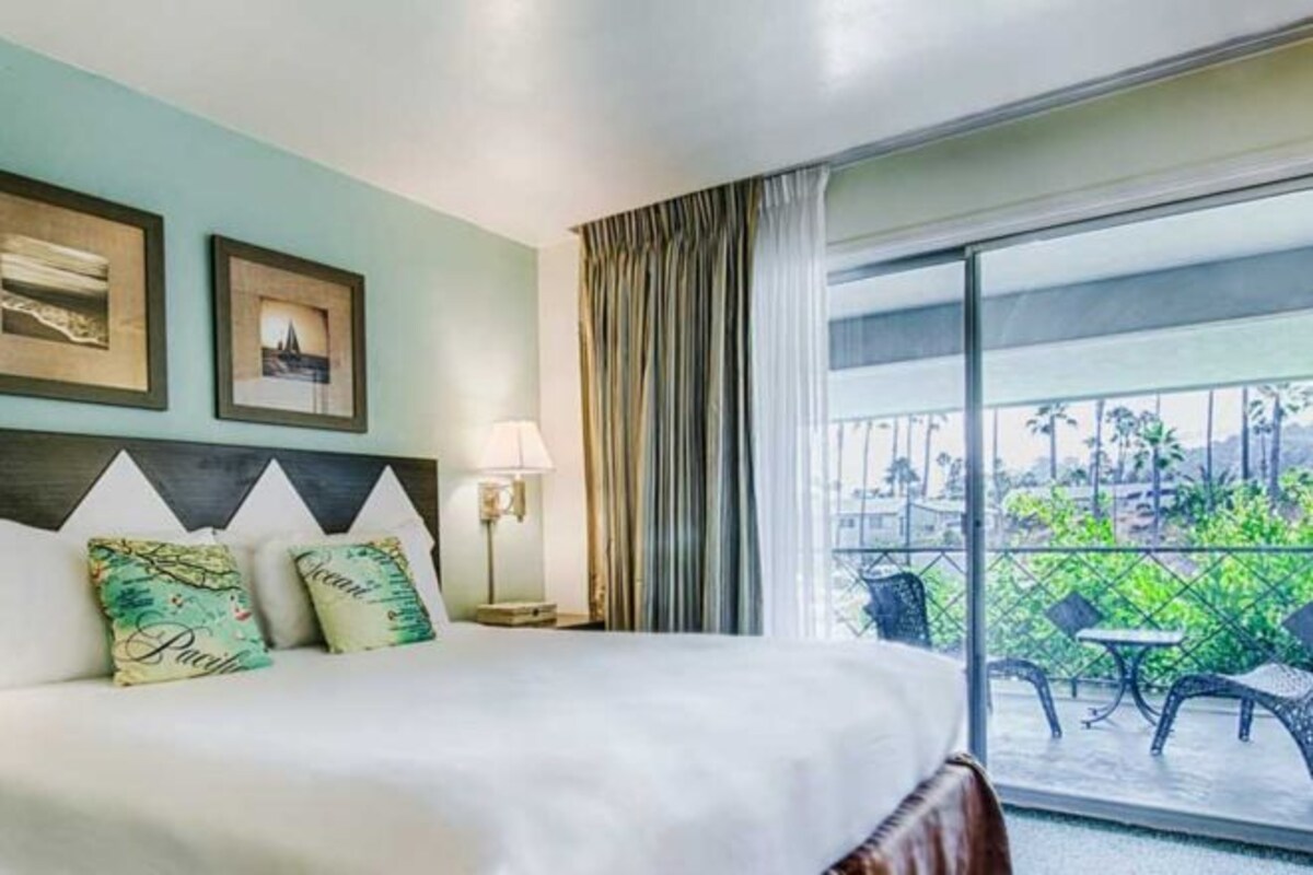 A hotel room with a bed, two framed pictures on the wall, a nightstand with a lamp, and a balcony with outdoor seating.