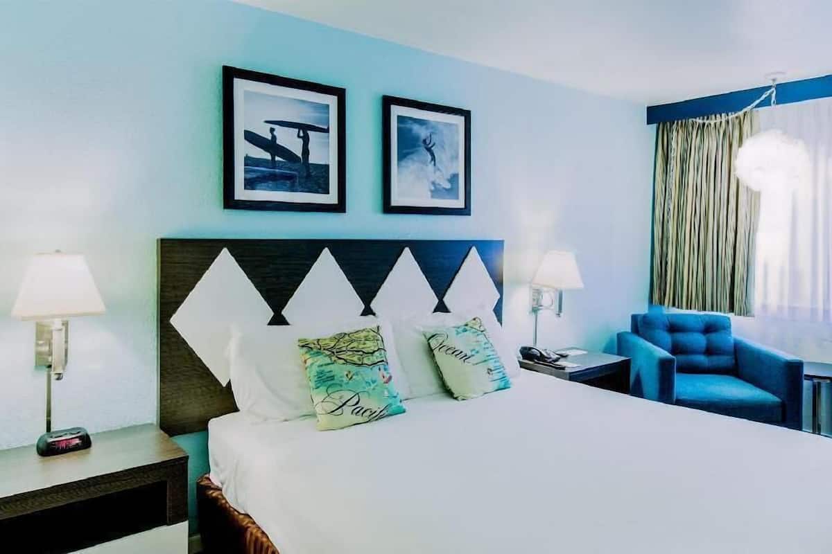 Quarto de hotel com cama, duas imagens emolduradas, uma cadeira azul e mesa de cabeceira com abajur.