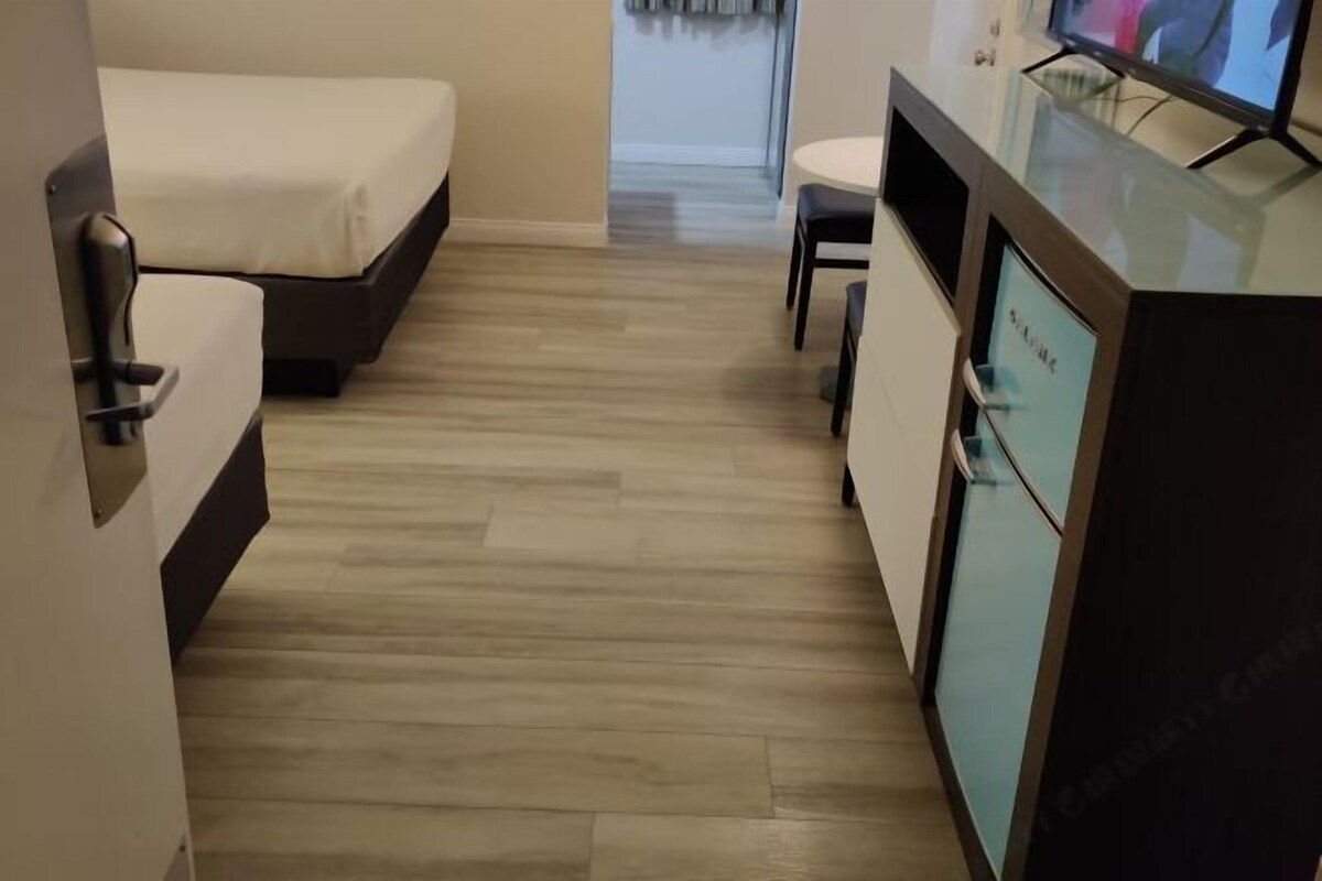 A compact hotel room with a bed, a small table, a TV, and a minibar.