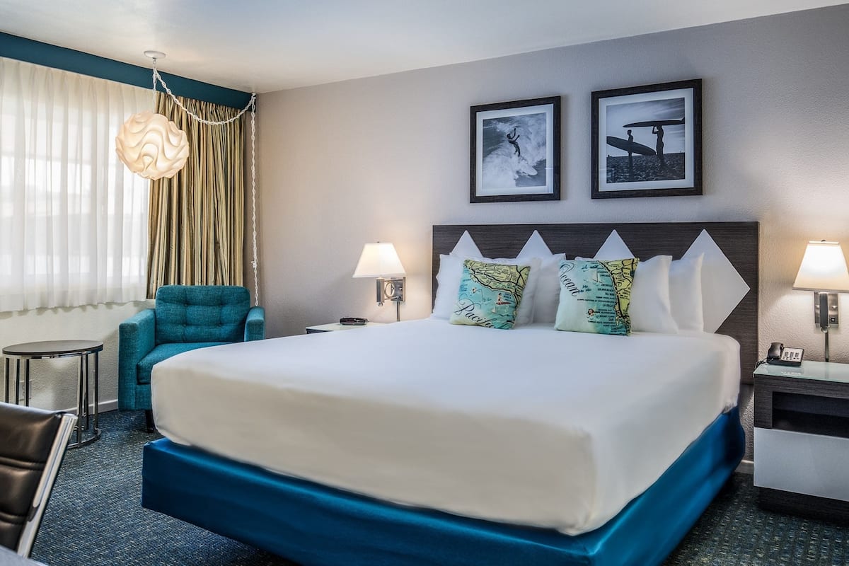 Quarto de hotel com uma cama grande, duas luminárias na parede, uma cadeira, mesa de cabeceira com telefone e uma janela com cortinas.