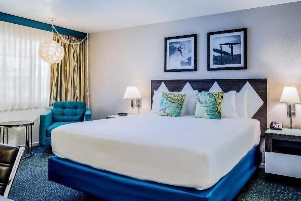 Quarto de hotel com uma cama grande, uma poltrona azul, duas luminárias fixadas na parede e duas molduras com quadros.