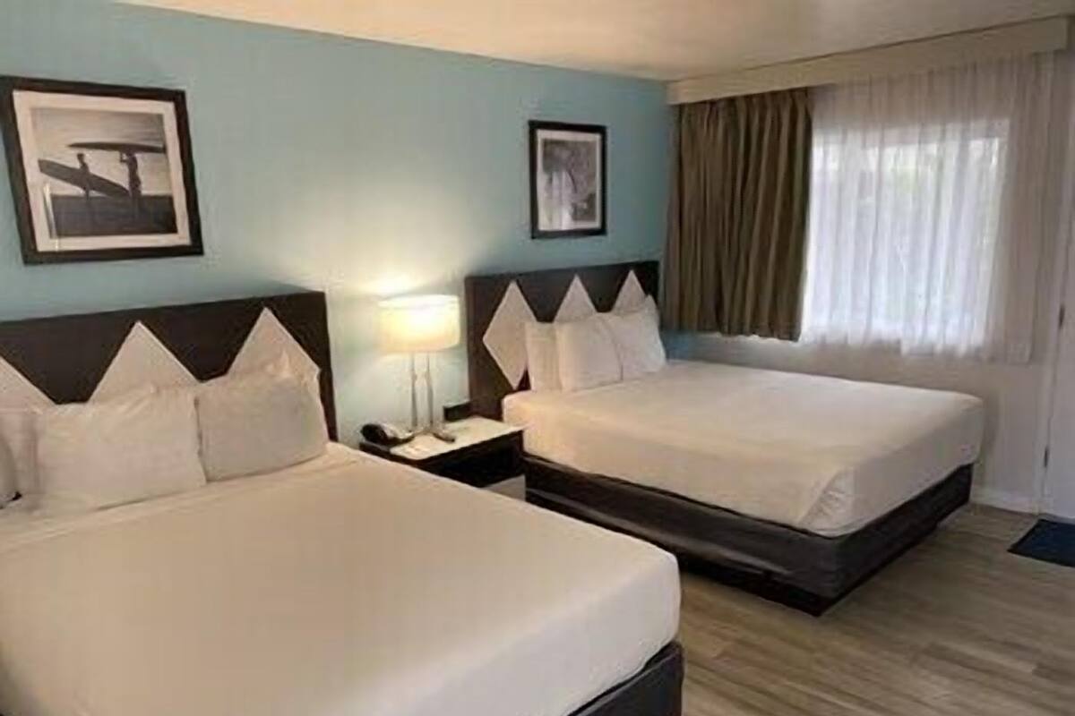 Quarto de hotel com duas camas, uma mesa de cabeceira com abajur e duas fotografias emolduradas na parede.