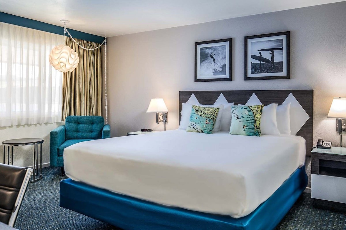 A hotel room with a large bed, two pillows with a map design, a blue accent chair, a small table, and a lamp on each side of the bed.