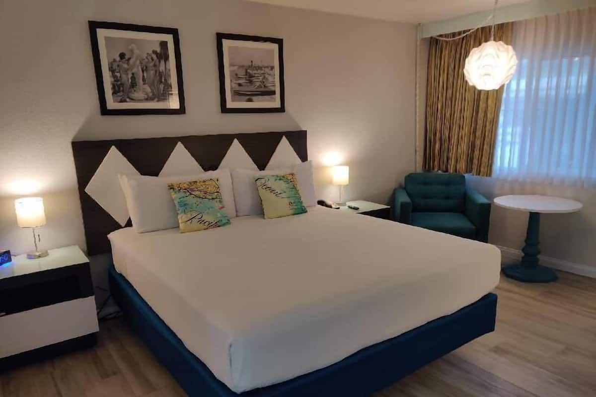 Quarto de hotel com uma cama, duas mesas de cabeceira, uma cadeira, uma mesinha e duas fotografias emolduradas na parede.