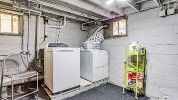 A laundry room with a washing machine, dryer, and a metal chair. There is a green cart with cleaning supplies.