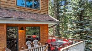 A wooden cabin with a deck, outdoor seating, and a large window overlooking a forest. - Aspen Brook Riversong by Estes Park Homes (Estes Park)