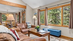 A spacious living room with leather sofas, a coffee table, and a blue bench. Large windows offer a view of trees. - Aspen Brook Riversong by Estes Park Homes (Estes Park)