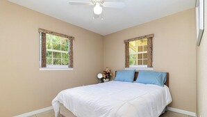 Ein Schlafzimmer mit einem Bett, zwei Vorhängen, einem Deckenventilator und einem kleinen Tisch mit Spiegel und Blumen.