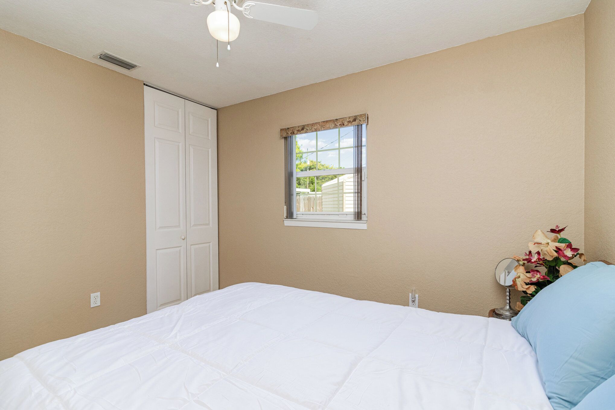 Une chambre à coucher avec un lit, une fenêtre, une table de chevet avec un miroir et un ventilateur de plafond.