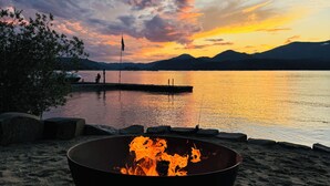 Une plage avec un foyer au premier plan, une jetée s’étendant dans une eau calme et des montagnes en arrière-plan.