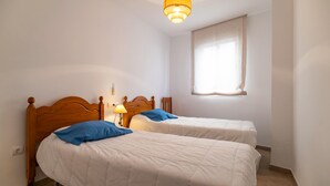 A room with two single beds, a nightstand with a lamp, a window with sheer curtains, and a wall-mounted light fixture.