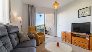 A modern living room with a grey sofa, a yellow armchair, a round coffee table, a wooden TV stand, and a large window with a view of the city.