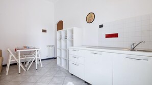 Una cuina compacta amb armaris blancs, un forn de mur, un rentat i una zona de dinar amb una cadira i taula.