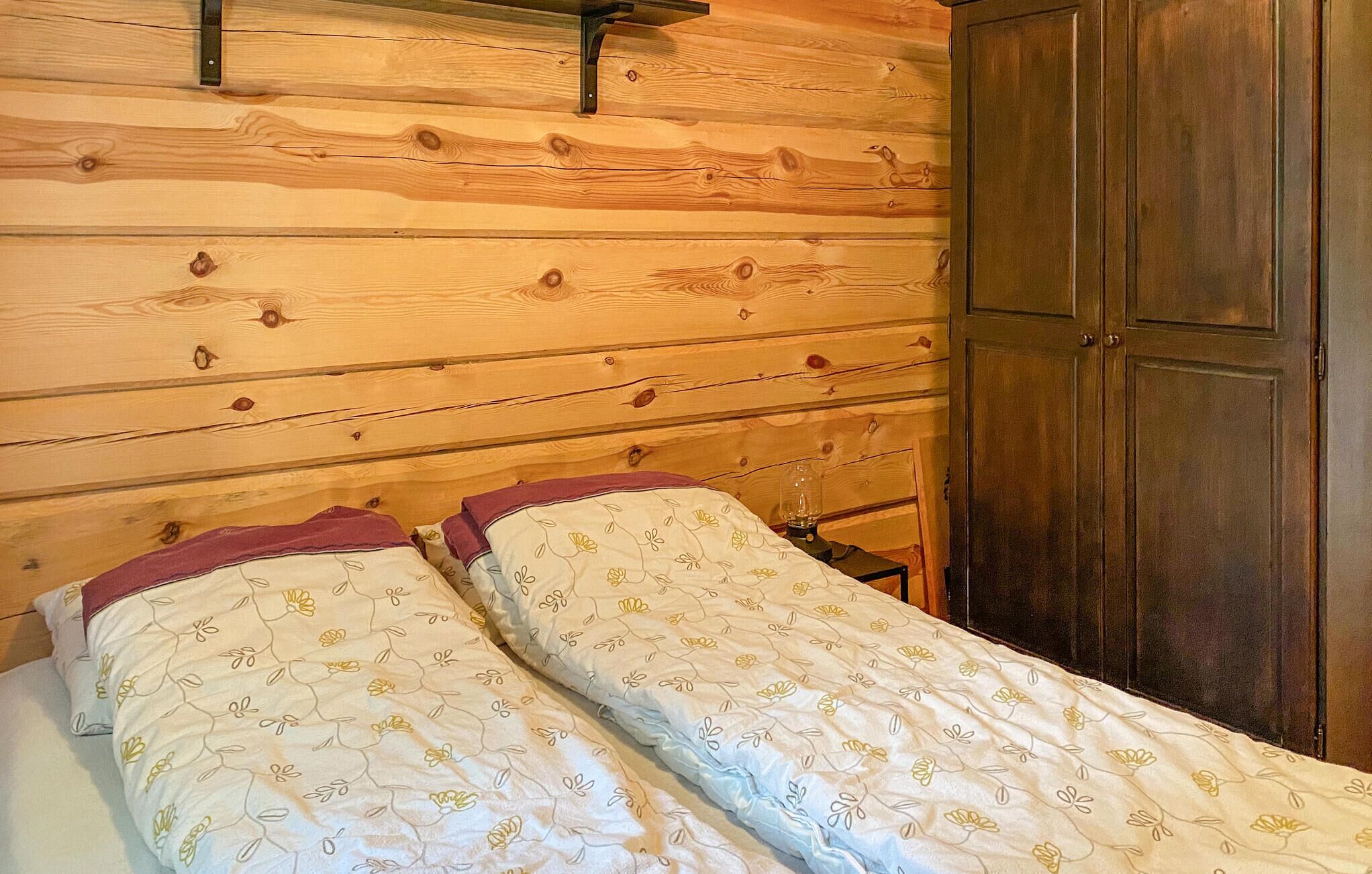 A wooden cabin room with two beds, a wooden wardrobe, and a small table with a lamp.
