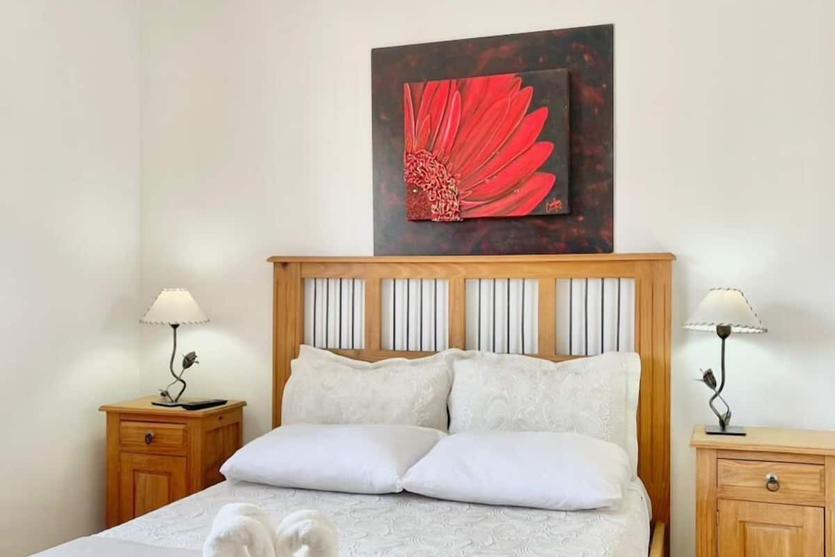 A neatly made bed with white linens, two bedside tables with lamps, and a large painting of a red flower on the wall.