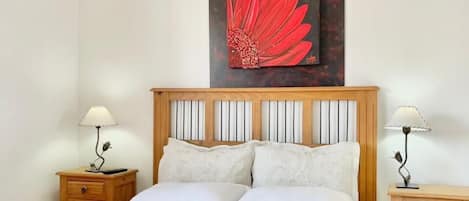 A neatly made bed with white linens, two bedside tables with lamps, and a large painting of a red flower on the wall.