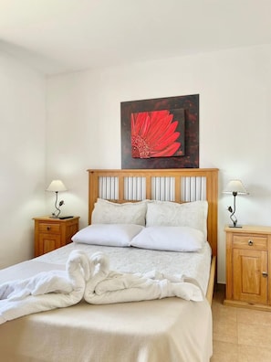 Una cama bien hecha con sábanas blancas, dos mesitas de noche con lámparas y un cuadro grande de una flor roja en la pared.