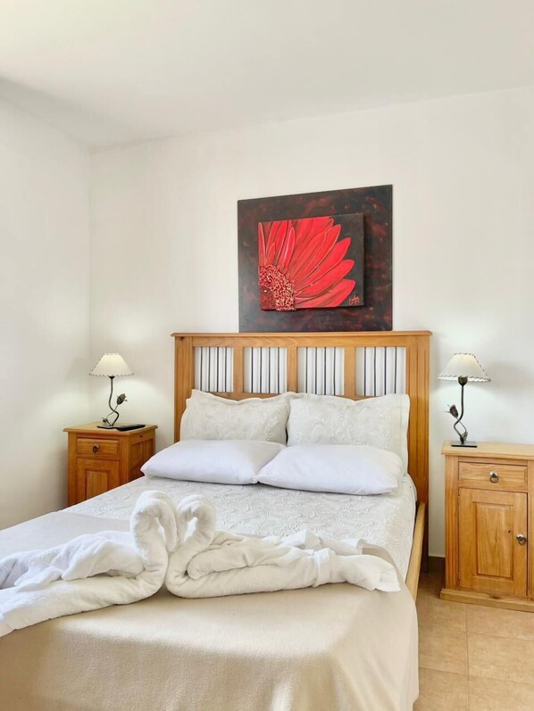 A neatly made bed with white linens, two bedside tables with lamps, and a large painting of a red flower on the wall.