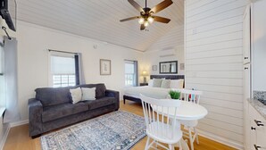 A room with a gray sofa, a white chair, a small table, a ceiling fan, and a bed with a headboard.