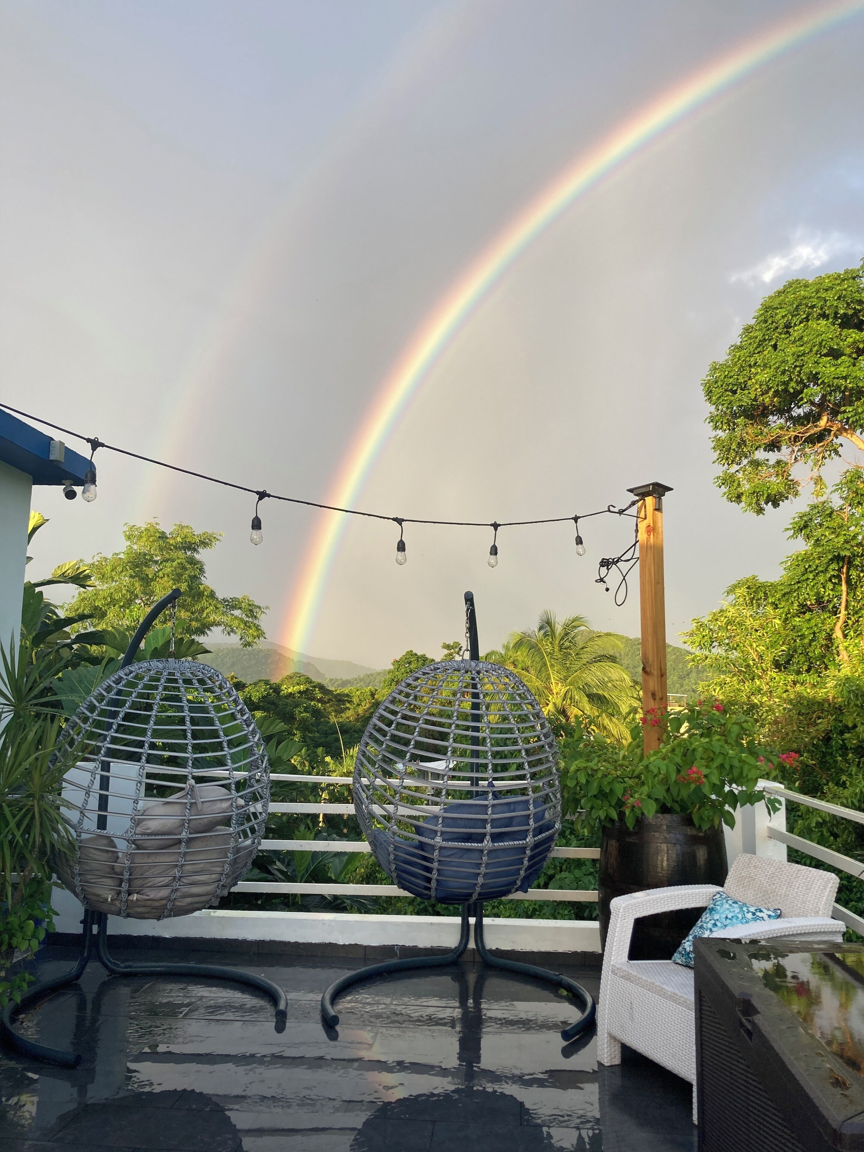 A rooftop terrace with two hanging egg chairs, a string of lights, and a view of a rainbow.