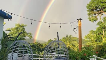 A rooftop terrace with two hanging egg chairs, a string of lights, and a view of a rainbow.