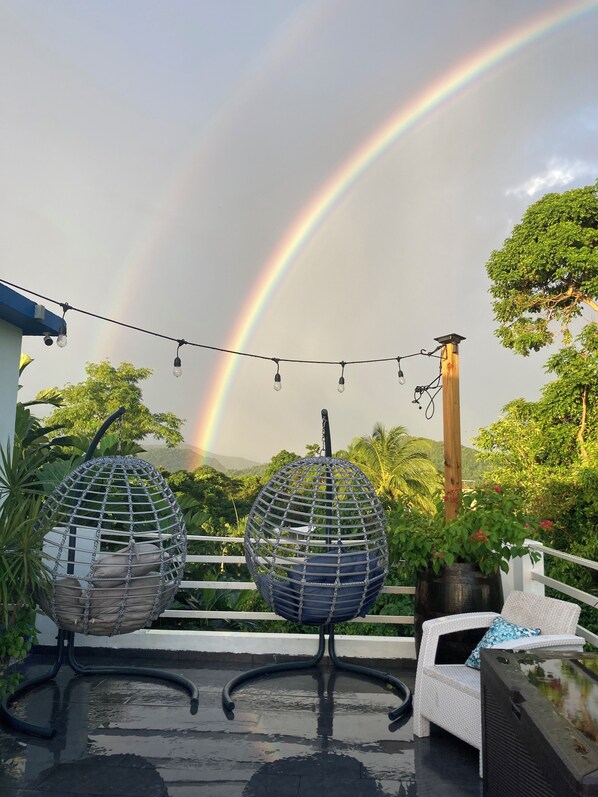 A rooftop terrace with two hanging egg chairs, a string of lights, and a view of a rainbow.