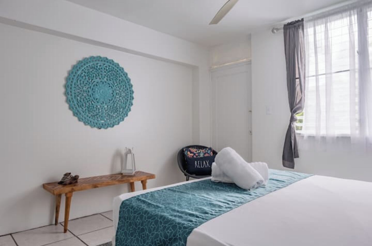 A hotel room with a bed, a small wooden table, a decorative wall hanging, and a window with curtains.