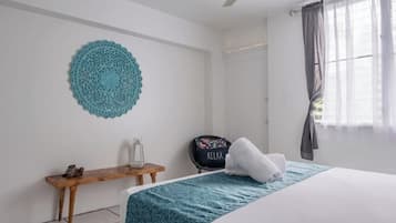 A hotel room with a bed, a small wooden table, a decorative wall hanging, and a window with curtains.