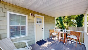 A covered porch with a wooden table and chairs, a door with a mailbox, and a white picket fence. - 1BR • King Bed • Sofa Bed • Sleeps 4 • POOL - 8 (Cape May)