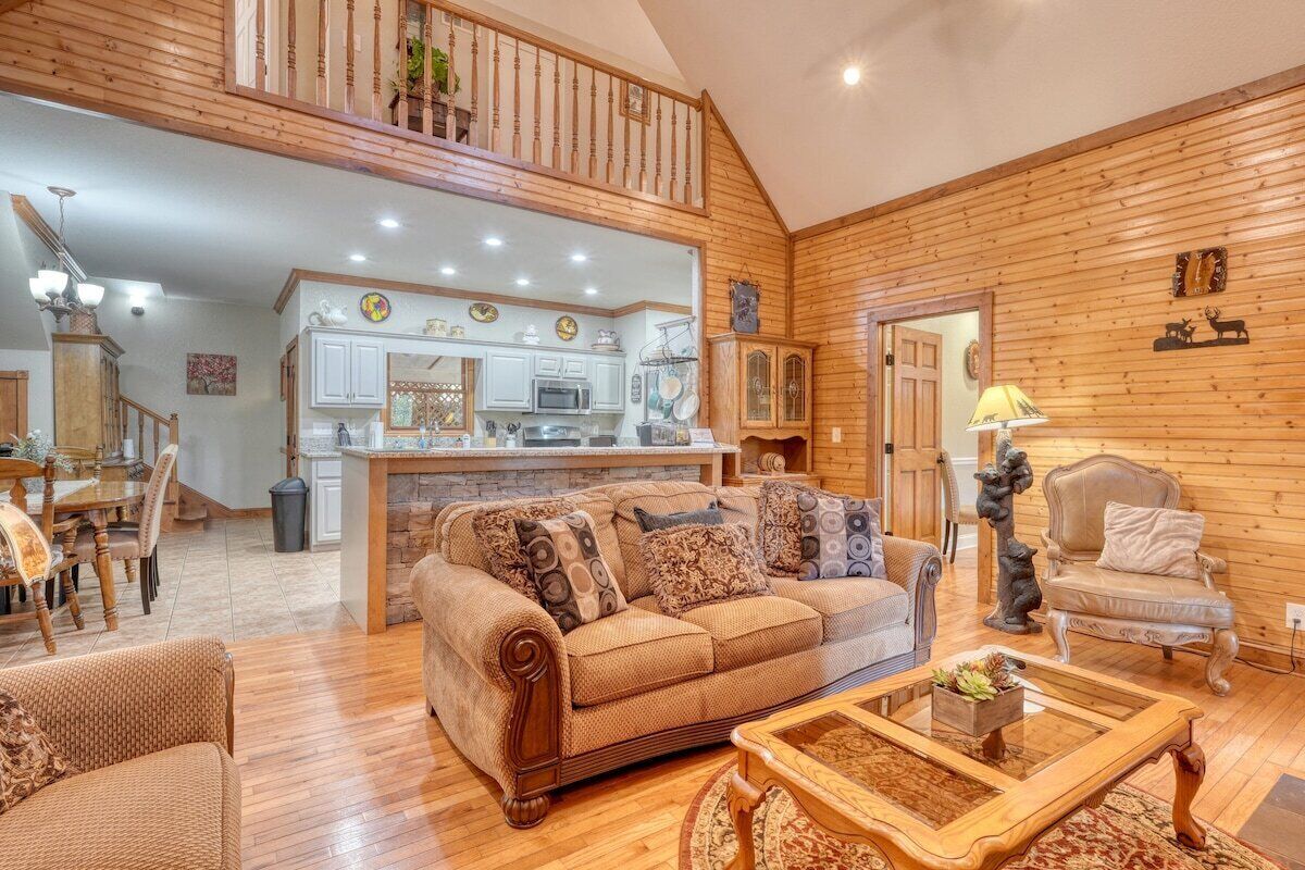 A cozy living room with a wooden ceiling, a sofa with patterned cushions, a coffee table, and a dining area with a chandelier.