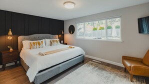 A bedroom with a large bed, a nightstand, a chair, a window with a view of greenery, and a ceiling light.