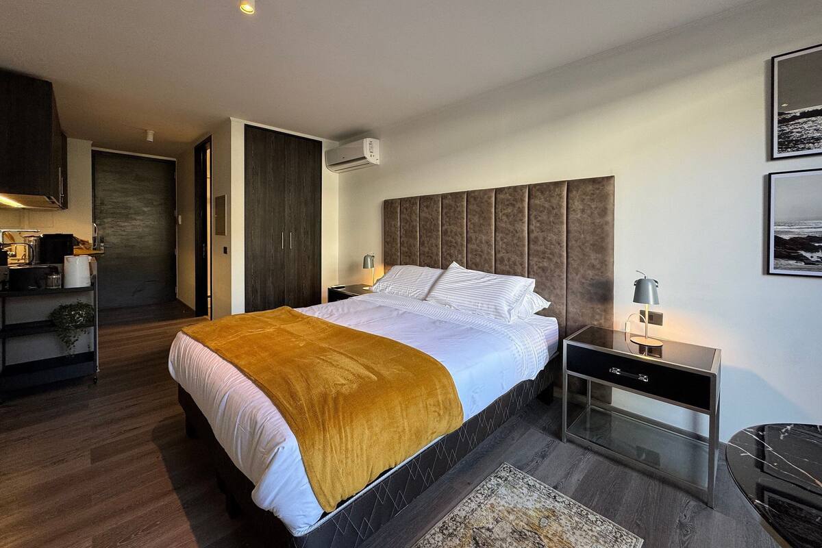Quarto moderno com uma cama grande, mesa de cabeceira, armário e cozinha ao fundo.