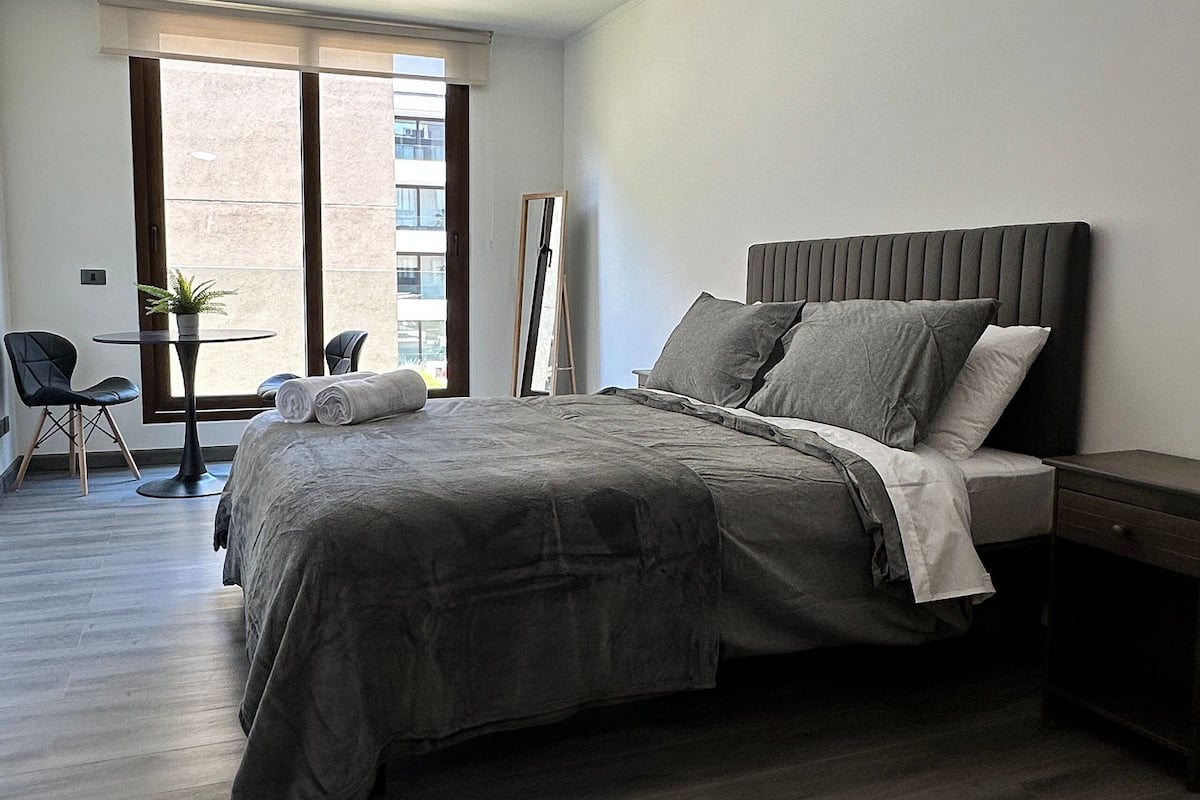 Quarto moderno com uma cama grande, uma mesinha de cabeceira, uma cadeira e uma janela com vista para prédios.