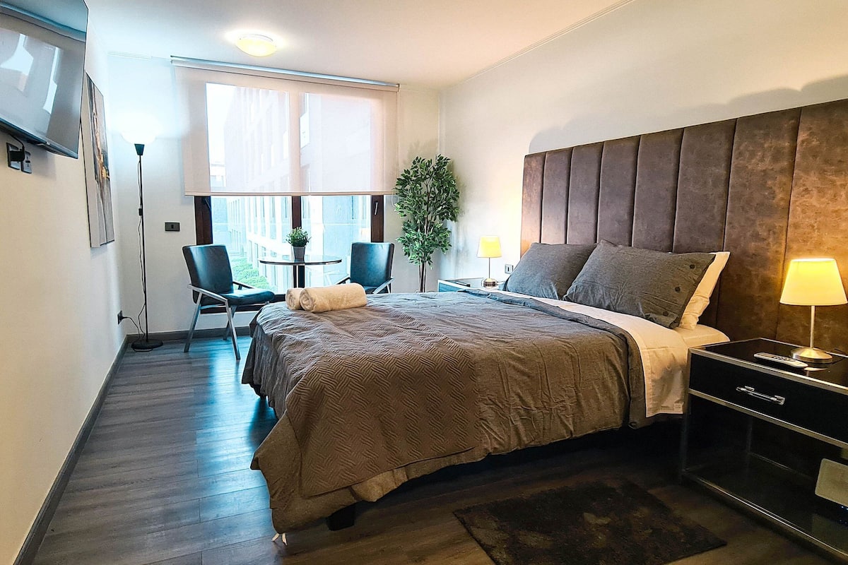 Quarto de hotel com cama, mesa de cabeceira, luminária, cadeira, mesinha e uma planta.