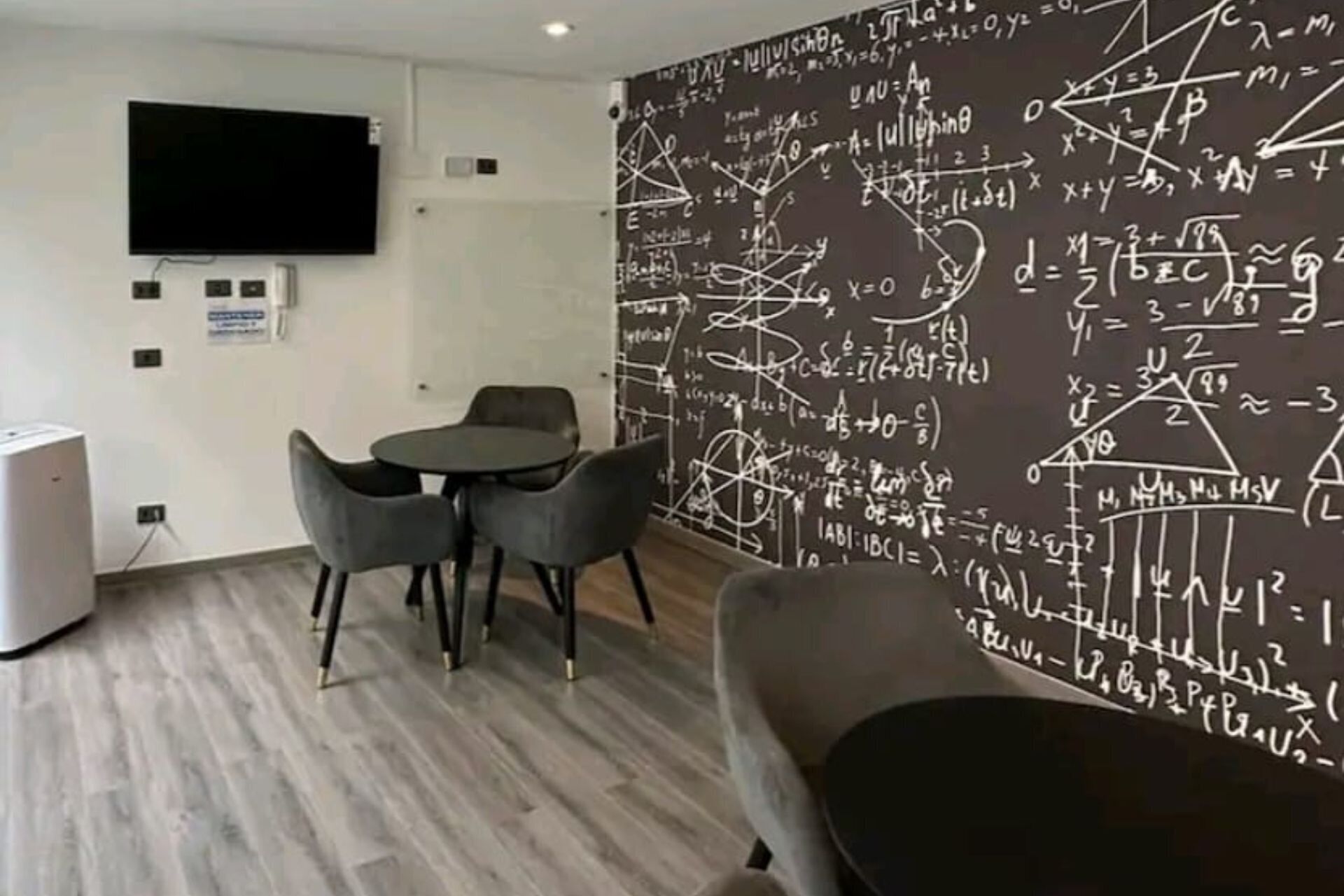 Una sala de reuniones moderna con una gran pizarra llena de ecuaciones y diagramas matemáticos, un televisor de pantalla plana montado en la pared y una mesa redonda con cuatro sillas.